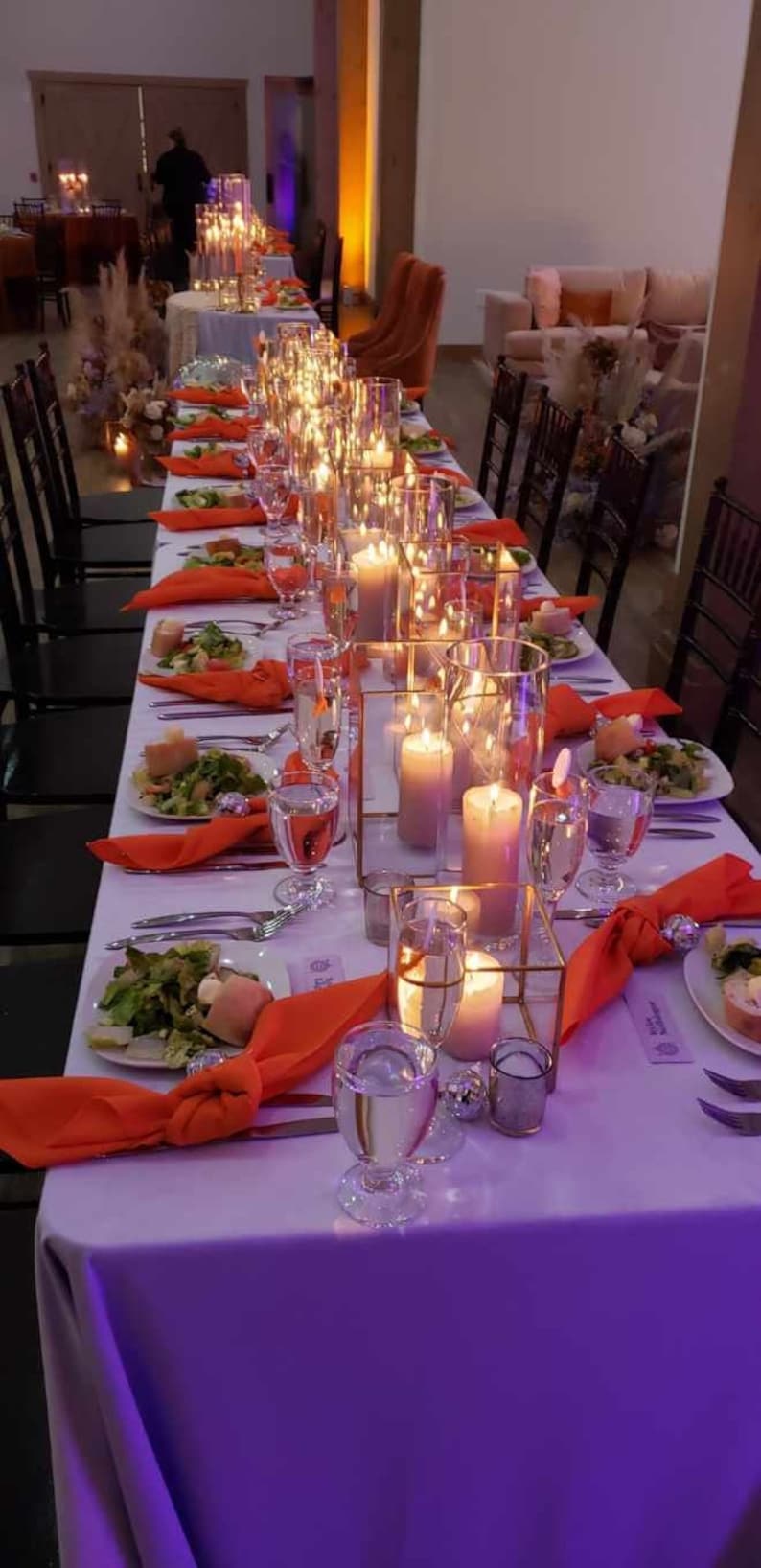 A long table set for a wedding reception with purple velvet table linens, orange napkins, and clear glass candle holders with lit candles. The table is decorated with greenery and flowers.