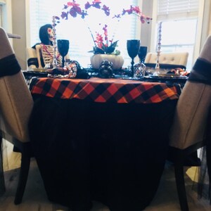 A Halloween table setting with a black and orange plaid tablecloth. The table is set with black plates, gold chargers, and black wine glasses. There are also black and white skull decorations on the table.