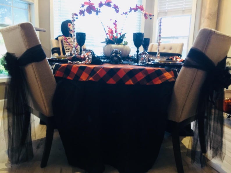 A Halloween table setting with a black and orange plaid tablecloth. The table is set with black plates, gold chargers, and black wine glasses. There are also black and white skull decorations on the table.