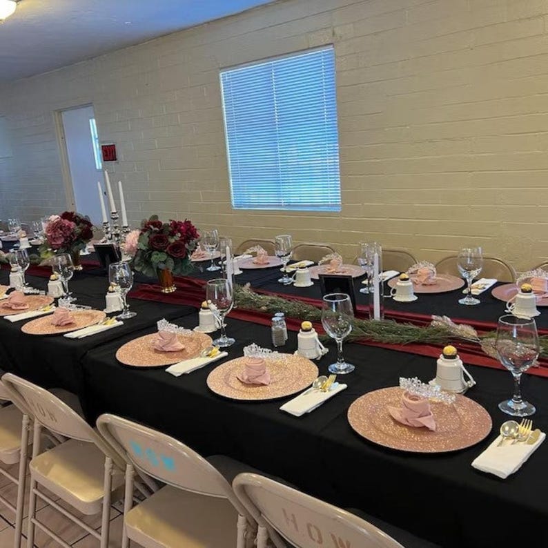 A burgundy velvet table runner drapes over a table with a black tablecloth set for a dinner party. There are velvet napkins on top of the dishes.