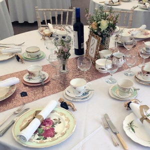 May include: A white table setting with a rose gold sequin table runner. The table is set with vintage china teacups, saucers, and plates. There are also gold animal figurines, white napkins, and wine glasses.