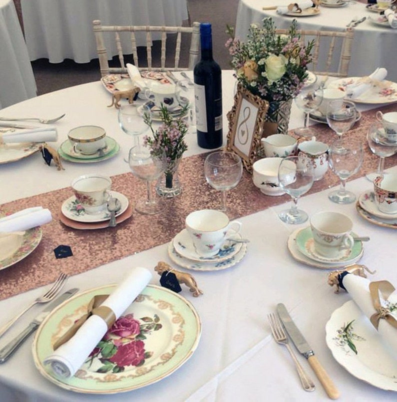 May include: A white table setting with a rose gold sequin table runner. The table is set with vintage china teacups, saucers, and plates. There are also gold animal figurines, white napkins, and wine glasses.