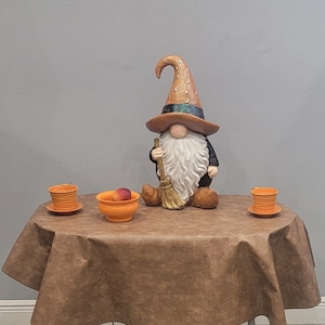 May include: A gnome figurine wearing a tall orange hat with moon and star patterns, a black band, and a long white beard. The gnome holds a broom. On the table are two orange cups on saucers and an orange bowl containing a single peach-colored fruit.