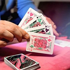 Puede incluir: Una mano sostiene cuatro naipes, cada uno con una sota de corazones, picas, tr&eacute;boles y diamantes. Las cartas tienen un fondo blanco con dise&ntilde;os gr&aacute;ficos en rosa y verde. Debajo hay una baraja con el reverso jaspeado en negro, rosa y verde.