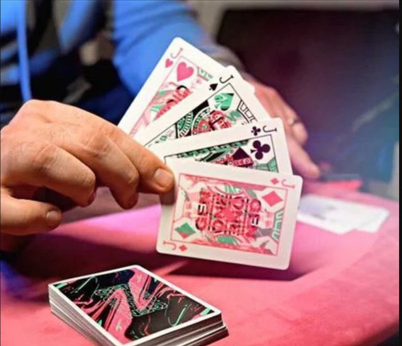 Puede incluir: Una mano sostiene cuatro naipes, cada uno con una sota de corazones, picas, tr&eacute;boles y diamantes. Las cartas tienen un fondo blanco con dise&ntilde;os gr&aacute;ficos en rosa y verde. Debajo hay una baraja con el reverso jaspeado en negro, rosa y verde.