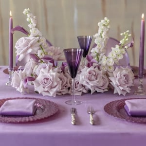 A table setting with a purple velvet tablecloth, purple plates, and purple velvet napkins. The centerpiece is a large arrangement of white and purple flowers with tall purple candles.