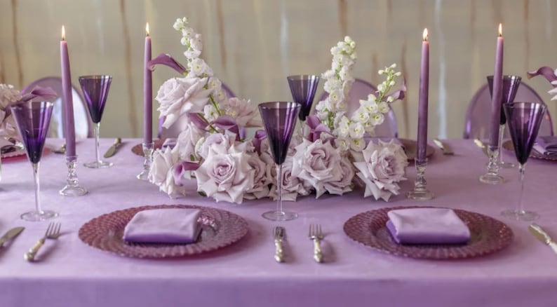 A table setting with a purple velvet tablecloth, purple plates, and purple velvet napkins. The centerpiece is a large arrangement of white and purple flowers with tall purple candles.