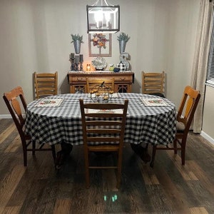 May include: A black and white checkered tablecloth covers a wooden dining table with six chairs. The table is set with placemats and a centerpiece of fall decor. The room has a light brown wood floor and white walls.