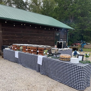 May include: A long buffet table covered with a black and white checkered tablecloth, featuring multiple wooden crates holding food containers, a silver metal pot, a stack of round pastries, and small floral arrangements in glass vases.