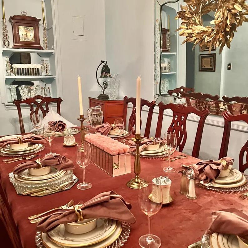 A blush velvet tablecloth with candlesticks in the center. The table is set with plates, blush velvet napkins, and clear glasses, set for an elegant meal at a home.