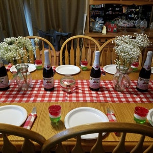 May include: A table setting with a red and white checkered tablecloth, four white plates, four clear glasses, four red and green layered desserts, and four bottles of sparkling wine. The table is set for a party or dinner.