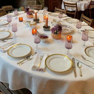 May include: A white tablecloth with a gold rimmed plate, silverware, and wine glasses set for a Thanksgiving dinner. There are candles, pumpkins, and flowers on the table.