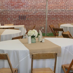 A rustic wedding reception tablescape with white tablecloths and burlap table runners. White flowers in mason jars are the centerpieces. Wooden folding chairs surround the tables.