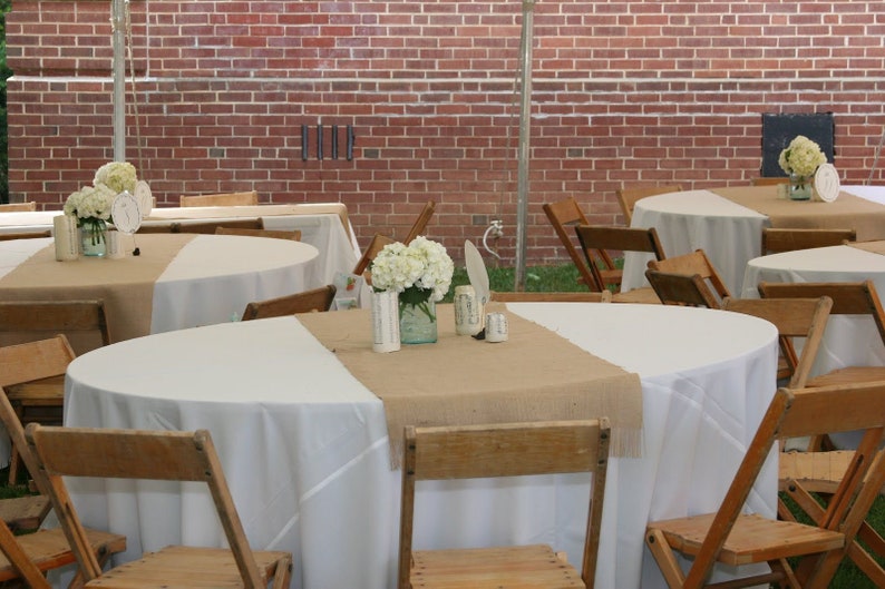 A rustic wedding reception tablescape with white tablecloths and burlap table runners. White flowers in mason jars are the centerpieces. Wooden folding chairs surround the tables.