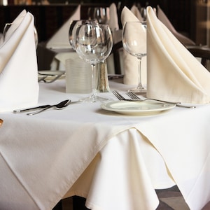 A restaurant table with a white tablecloth is set with white napkins folded into triangles, silverware, and clear glass wine glasses. The table is set for a formal dinner.