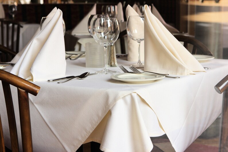 A restaurant table with a white tablecloth is set with white napkins folded into triangles, silverware, and clear glass wine glasses. The table is set for a formal dinner.