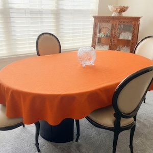 May include: An oval dining table covered with a solid orange tablecloth. The table is surrounded by four chairs with dark wood frames and cream-colored upholstered seats. A clear, textured glass bowl sits in the center of the table.