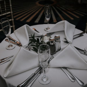 A white tablecloth with four place settings, each with a white dinner napkin folded into a triangle, a silver fork, a silver knife, and a champagne flute. There are two salt and pepper shakers in the center of the table, along with a sprig of greenery.