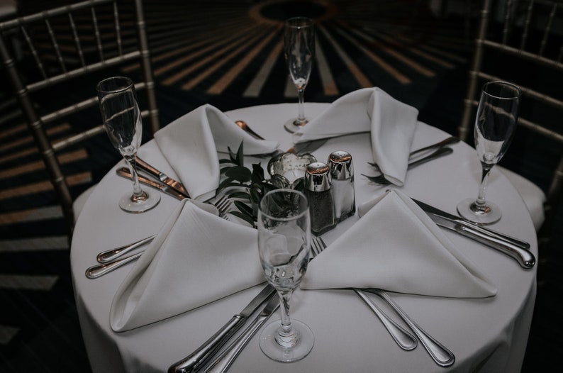A white tablecloth with four place settings, each with a white dinner napkin folded into a triangle, a silver fork, a silver knife, and a champagne flute. There are two salt and pepper shakers in the center of the table, along with a sprig of greenery.