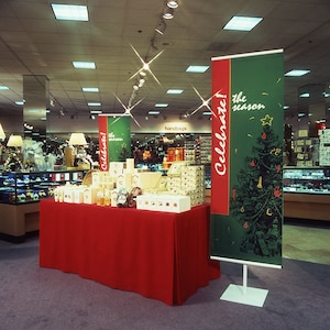 May include: A red table covered in a red tablecloth with various gift boxes and candles on top. Two green and red banners with the text "Celebrate! the season" stand behind the table.