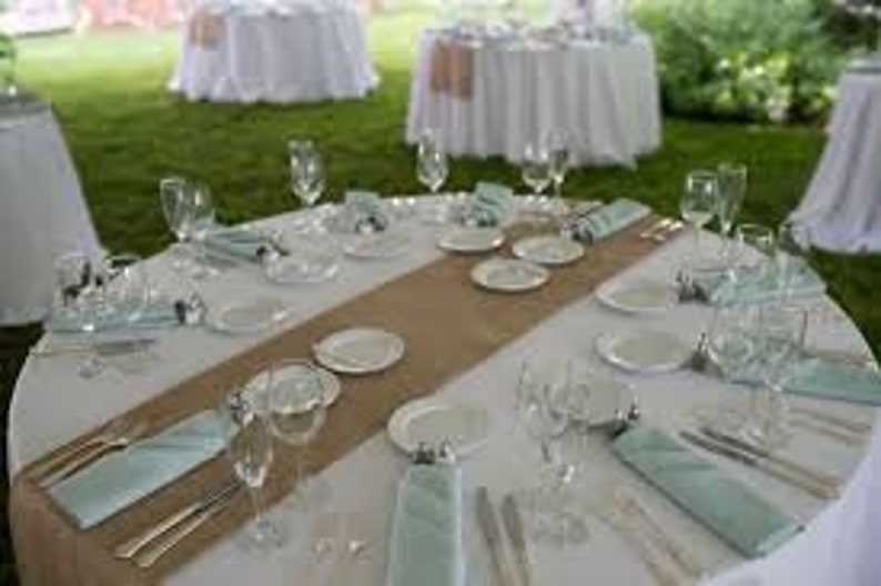 A round table set for a wedding reception with white linens, light blue napkins, and a burlap runner down the center.