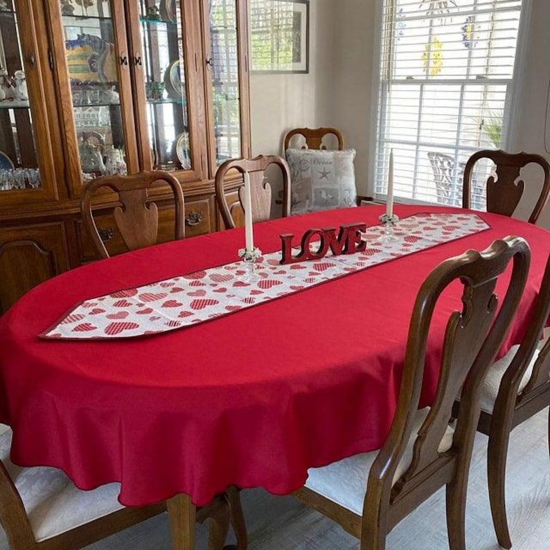 An oval table in the dining room of a home with a red tablecloth and white table runner. Candles and a love decoration are on the romantic table setting.