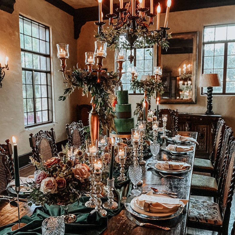 A formal dining table setting with a green velvet tablecloth, gold candelabras, and a three-tiered green cake. The table is set with white plates, gold silverware, and crystal glasses. The room is decorated with greenery and candles.