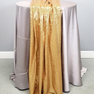 May include: A gold sequin table runner draped over a light gray tablecloth. The runner is made of a soft, shimmering fabric with a subtle sheen.