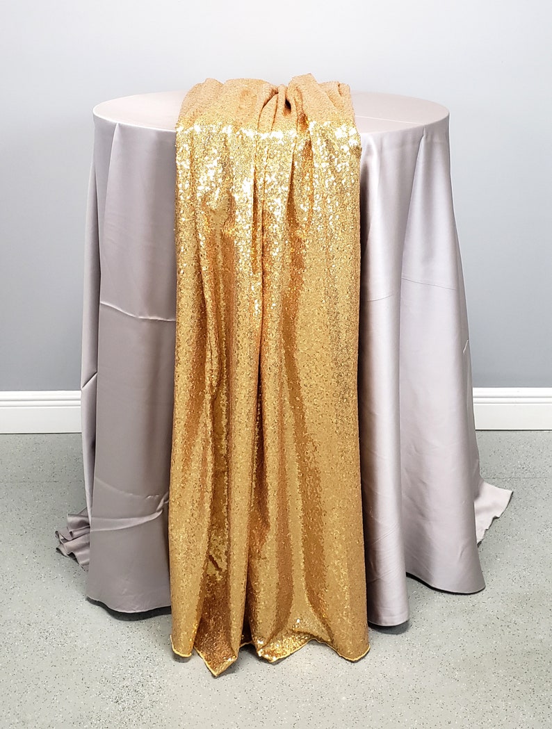 May include: A gold sequin table runner draped over a light gray tablecloth. The runner is made of a soft, shimmering fabric with a subtle sheen.