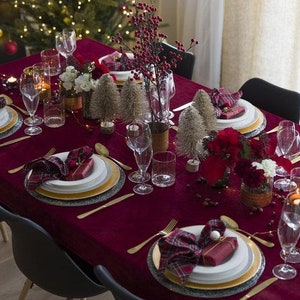 A burgundy velvet tablecloth  and velvet napkins with gold chargers and white plates. There are small silver trees, red berries, and red flowers as centerpieces. The table is set for a holiday dinner.