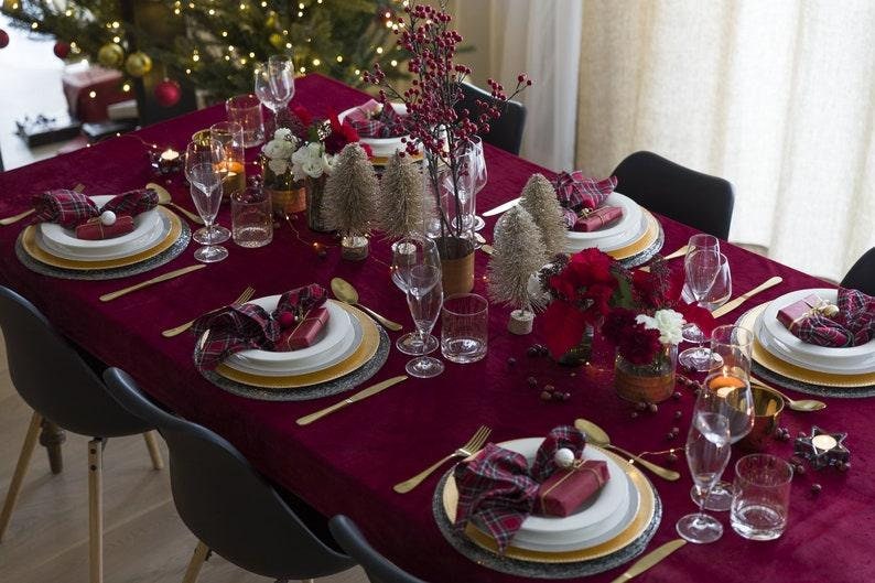 A burgundy velvet tablecloth  and velvet napkins with gold chargers and white plates. There are small silver trees, red berries, and red flowers as centerpieces. The table is set for a holiday dinner.