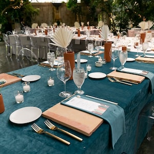 A teal velvet tablecloth and velvet napkins with white plates, gold silverware, and wooden chargers. There are clear glass water glasses and small white candles on the table. The table is set for a formal dinner.