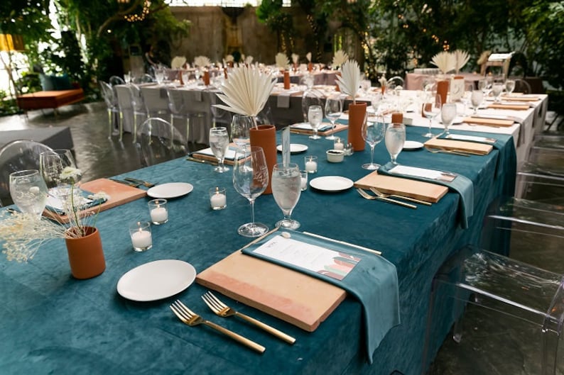 A teal velvet tablecloth and velvet napkins with white plates, gold silverware, and wooden chargers. There are clear glass water glasses and small white candles on the table. The table is set for a formal dinner.
