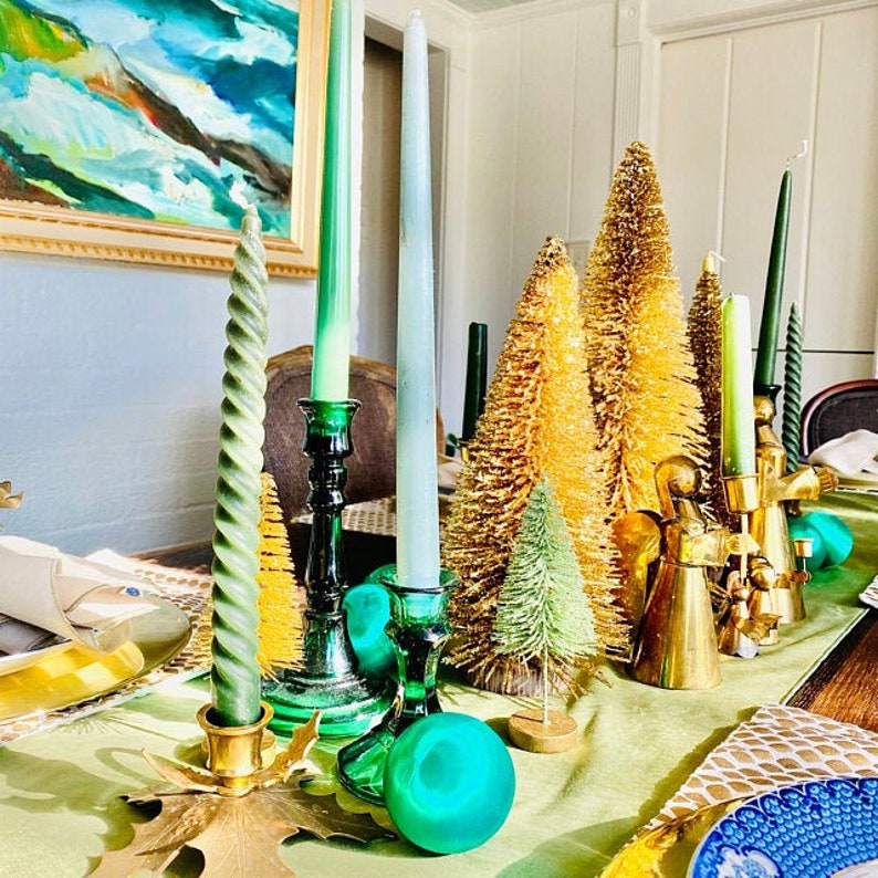 May include: A table setting with green and gold candlesticks, green and blue candles, and gold and green Christmas trees. The table is covered in a green tablecloth.