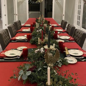 A large oblong table with a red tablecloth, hunter green velvet table runners and red embroidered napkins set for a holiday dinner.