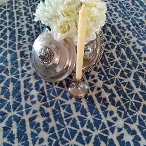 A silver serving dish with a lid, a silver candlestick holding a cream-colored candle, and a bouquet of white flowers sit on a blue and white Shibori round tablecloth.