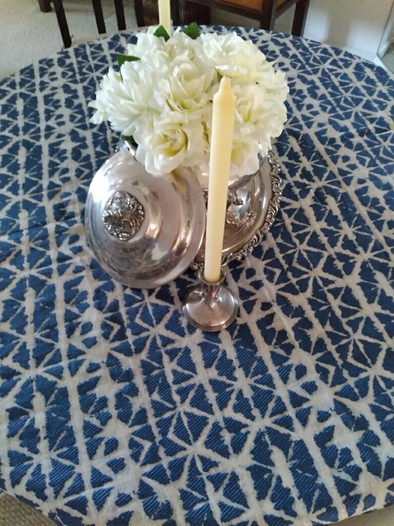 A silver serving dish with a lid, a silver candlestick holding a cream-colored candle, and a bouquet of white flowers sit on a blue and white Shibori round tablecloth.