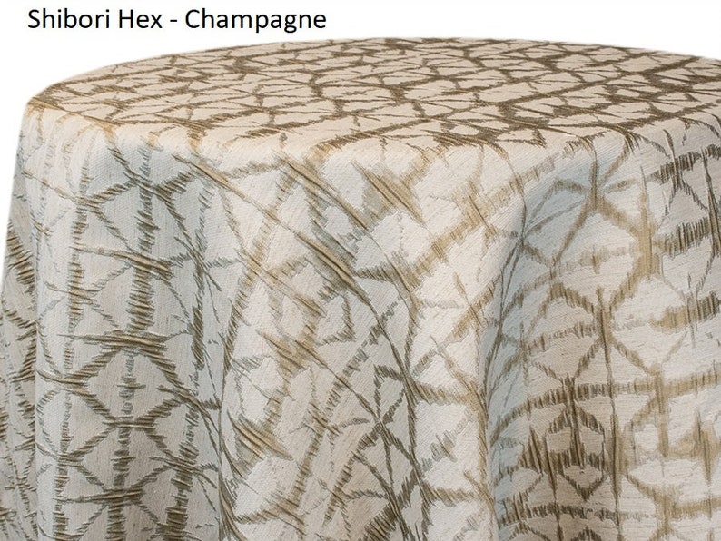 Champagne Shibori tablecloth with a geometric Shibori Hex pattern. The fabric has a textured appearance with a repeating diamond and line design. The tablecloth is draped over a round surface. The tablecloth manufacturer is Premier Linens.