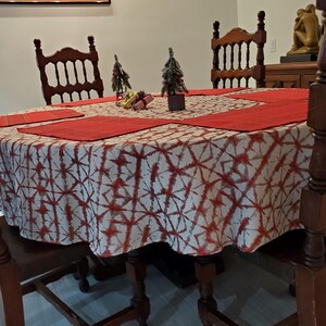 May include: A round dining table set for a holiday meal. The table is covered with a white and red patterned tablecloth and red placemats. Two small Christmas trees and decorative gifts are on the table. Wooden chairs surround the table.