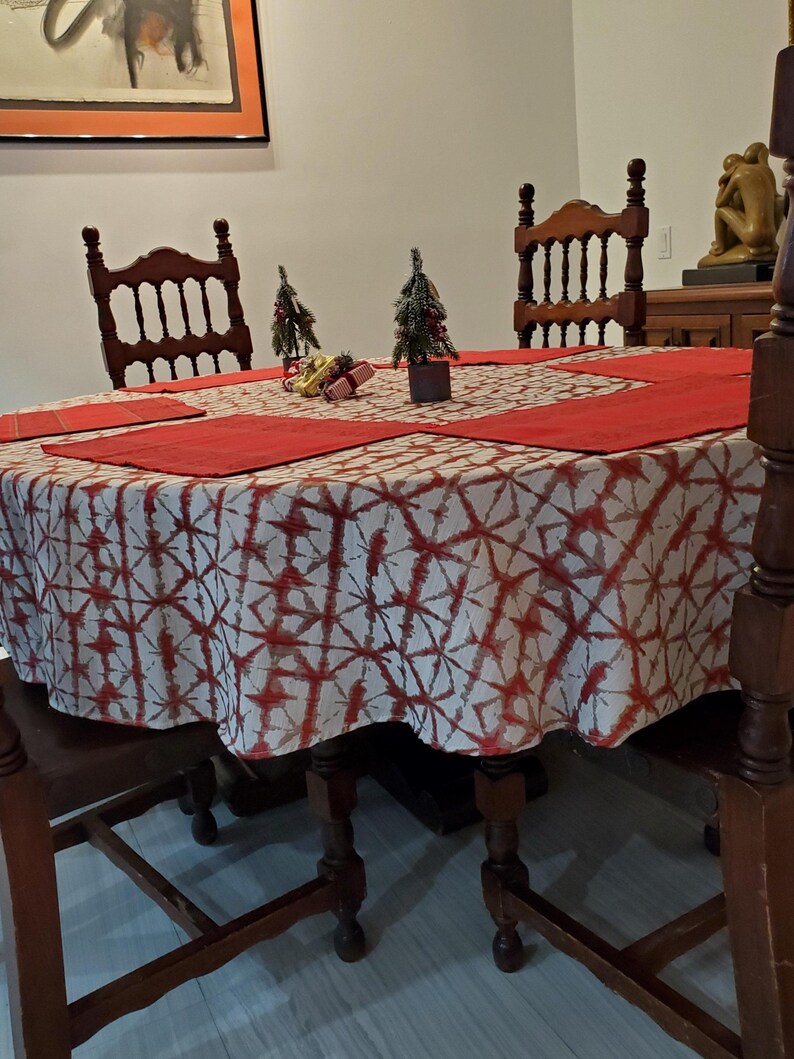May include: A round dining table set for a holiday meal. The table is covered with a white and red patterned tablecloth and red placemats. Two small Christmas trees and decorative gifts are on the table. Wooden chairs surround the table.