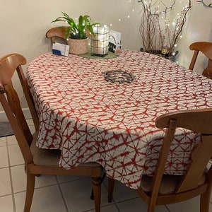 An oval tablecloth, the fabric is a red and white Shibori tablecloth. Four wooden chairs surround the table. A painting of apples hangs on the wall above the table. A potted plant and a decorative candle holder are on the table.