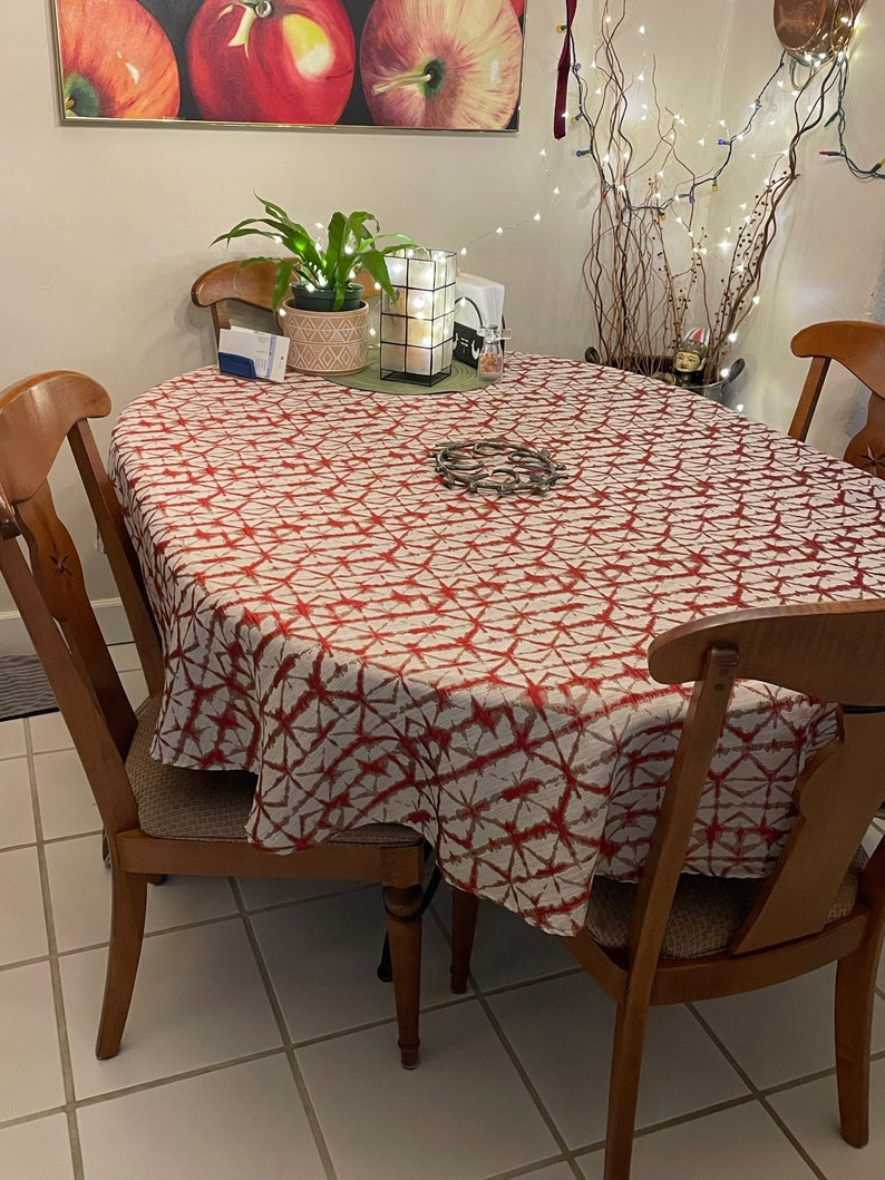 An oval tablecloth, the fabric is a red and white Shibori tablecloth. Four wooden chairs surround the table. A painting of apples hangs on the wall above the table. A potted plant and a decorative candle holder are on the table.
