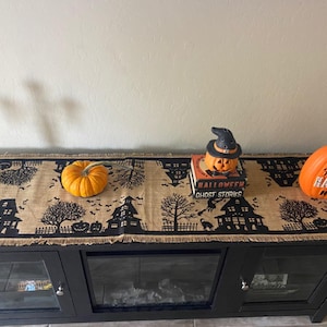 A black and brown Halloween table runner with a frayed edge, featuring black silhouettes of houses, bats, and trees. The runner is decorated with a small orange pumpkin, a jack-o'-lantern with a witch's hat, and a larger orange pumpkin with the text "Home Sweet Haunted Home".