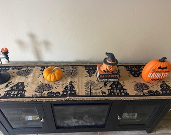 May include: A black and brown Halloween table runner with a frayed edge, featuring black silhouettes of houses, bats, and trees. The runner is decorated with a small orange pumpkin, a jack-o'-lantern with a witch's hat, and a larger orange pumpkin with the text "Home Sweet Haunted Home".
