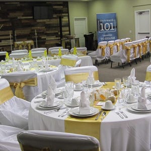 A banquet hall with many tables with white tablecloths and white napkin formally set for an event. The dinner napkins are cotton and they are in a standing fold on top of the plates.