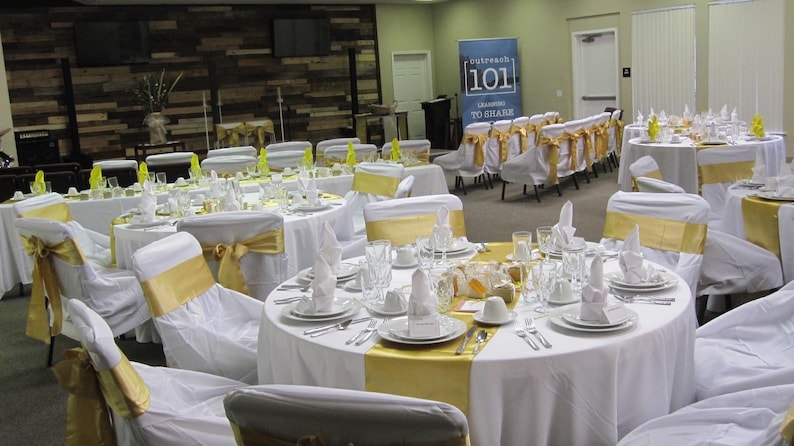 A banquet hall with many tables with white tablecloths and white napkin formally set for an event. The dinner napkins are cotton and they are in a standing fold on top of the plates.