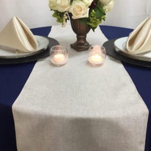 May include: A table setting with a navy blue tablecloth, a cream-colored linen table runner, and white plates with folded napkins. Two small glass candle holders with lit candles and a floral arrangement in a bronze vase are centered.