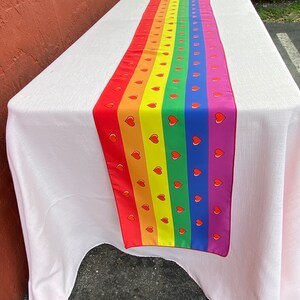 May include: A rainbow table runner with red hearts on each stripe. The runner is on a white tablecloth.
