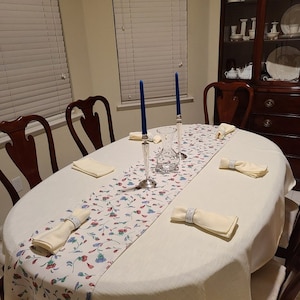 May include: A white tablecloth with a floral runner and four place settings. Each place setting includes a white napkin with a ring and a blue candle. The table is set for a formal dinner.
