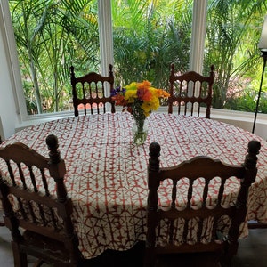A oval wooden dining table set with six chairs. The table is covered with a Shibori tablecloth in red and white. A vase of colorful flowers sits in the center. The chairs are made of dark wood.